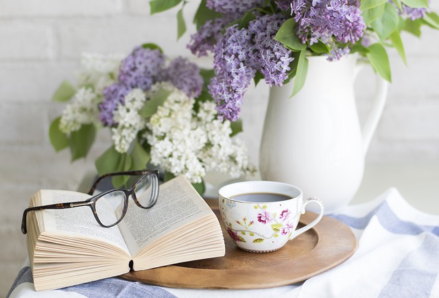 Photo of an open book with glasses on it next to a tea cup and a white pottery jug with small flowers